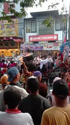 A crowd of people are gathered on a street, watching a performance. There are shops lining the street, including "SONIC CHICKEN" and "MOMOYO". Performers in elaborate costumes are dancing and moving in the street. Some of the costumes appear to be horse-like. Many people in the crowd are holding up their phones, presumably to record the event. The overall atmosphere appears festive.