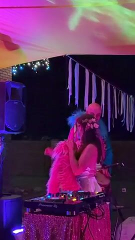 Two people are positioned in front of a DJ setup on a table covered with a pink sequined cloth. The person on the left, appearing to be a woman, is wearing a white dress and a fluffy pink jacket, with a flower crown in her hair. She is leaning into the person on the right. The person on the right, appearing to be a man, is wearing a colorful, fuzzy jacket and is playing a drum. They are both under a colorful canopy illuminated with pink and yellow lights. In the background, there is a speaker, some string lights, and a brick wall. The scene appears to be outdoors at night.