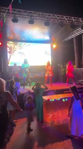 A group of children in colorful, tropical-themed outfits are dancing on a stage. Behind them, a large screen displays an image of palm trees and a beach. The stage is illuminated by colored lights, primarily pink and blue, and is set up outdoors, likely at night, as indicated by the darkness of the background and the artificial lighting. There is an audience of adults and children watching the performance from the foreground. The overall atmosphere appears to be festive and lively, possibly a luau or a similar themed event.