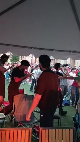 The video shows a group of people gathered under a large tent, possibly for an event or celebration. The lighting suggests it is late afternoon or early evening. In the foreground, several folding chairs and tables are visible, some with orange and red accents. In the middle ground, a group of young men are interacting. One man in a maroon shirt faces away from the camera, with his hands behind his head. To his left, a man in a black t-shirt is looking at his phone. To the right of the man in the maroon shirt, another man in a green and yellow tie-dye shirt is gesturing with his arms outstretched. Further back, a crowd of people are standing and watching. Many are wearing red and green clothing, suggesting a possible affiliation or theme. Some people are holding flags, also in red and green. The background shows a building with arched windows and columns. The ground appears to be covered in artificial turf. The overall atmosphere seems lively and social.