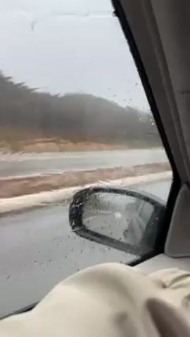 The video shows a view from inside a car, looking out the passenger side window. It is raining heavily, with numerous raindrops visible on the window. The windshield wipers are active. Outside, a road is visible, wet from the rain, with some orange construction barriers along the side.  Across the road is a blurry view of trees and vegetation. The car's side mirror is visible. A portion of what appears to be a light-colored jacket or sleeve is visible near the bottom of the frame.
