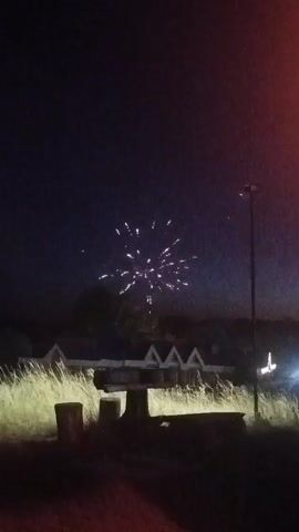 The video shows a nighttime scene with a fireworks display. In the foreground, there is a picnic table and some concrete structures. Beyond the table, there are some houses with triangular roofs. A single firework explodes in the dark sky, illuminating the scene with its bright light. A street lamp is visible to the right of the frame.