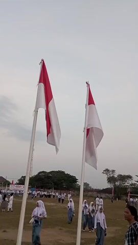 The video shows an outdoor scene with two Indonesian flags flying on poles in the foreground. The flags are red and white, with the red portion on top. The flags are draped, suggesting a slight breeze. In the background, a group of people, mostly young individuals dressed in white shirts and some wearing hijabs, are gathered in an open grassy field. Some of these individuals appear to be walking or standing in formation. There are trees and buildings visible in the distance. The sky is overcast and appears to be late afternoon or early evening. The overall impression is of a school event or ceremony taking place.
