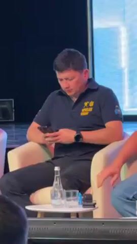 Here is a description of the video:
The video shows a man in a navy polo shirt with the words "MYKOLAYIV REGION" on it sitting in a white chair on a stage. He is looking at and using a smartphone. He is wearing a watch on his left wrist. To his left is a small white table with a bottle of water and a glass on it, as well as a small microphone. The man is sitting next to another person, only their arm and part of their shoulder are visible. The background is a blue screen.