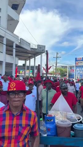 The video shows a crowd of people marching in a street, many wearing red shirts and carrying red flags. They appear to be participating in a demonstration or political rally. Some people are carrying water bottles. In the foreground, a street vendor's cart covered in a transparent plastic sheet can be seen. The environment appears to be a city street, lined with buildings and utility poles. The sky is partly cloudy.