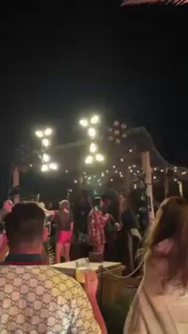 The video shows an outdoor party at night. A band is performing on a stage with multiple spotlights. People in the crowd are dancing and drinking. Palm trees are visible around the perimeter, also lit up. Some attendees are taking pictures or recording the event on their phones.