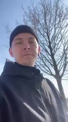 A man films himself in front of a snow-covered golf course on a sunny day. He is wearing a black beanie and a dark-colored jacket. He is standing on a paved path. In the background, the golf course is covered in snow with some patches of green visible. There are trees in the distance. The sky is clear and blue.