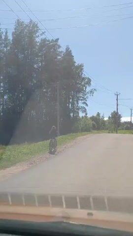 A person rides a bicycle on a paved road next to a grassy shoulder. They are dressed in dark clothing and a helmet. Tall trees line the left side of the road, and power lines are visible overhead. The sky is clear and blue. The video appears to be filmed from inside a vehicle.