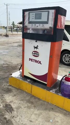 The video shows a petrol station with an MRS Petrol pump on a yellow-tiled platform. Beside it are a fire extinguisher in a purple basket and a red bucket labeled "Fire Bucket Filled With Sand". A white van is parked nearby and a man stands facing another petrol station labeled "ARIGBABU OIL & GAS" across the open space. People and vehicles, including motorcycles and yellow vans, are moving around in the distance. The sky is overcast.