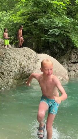 In a sun-dappled, lush green forest setting, three young boys are enjoying a refreshing dip in a clear, turquoise river. The scene unfolds near Khleborob, Russia, under what appears to be pleasant weather, possibly late morning or early afternoon, given the bright lighting.

The foremost subject is a fair-haired boy in teal swim trunks, mid-leap into the water. His face is turned towards the camera, a slight grimace of anticipation or exertion visible as he splashes into the river. Just behind him, another boy, also blond and in dark swim trunks, is already in the water, swimming with his arms outstretched in a freestyle stroke, his expression determined. Further back, perched on a large, moss-covered rock, a third boy in light-colored swim trunks stands poised, looking down into the water, about to join his friends.

The river itself is a vibrant aquamarine, reflecting the surrounding greenery and the large, textured rocks that line its banks. Towering trees with dense foliage form a verdant canopy overhead, creating a natural, secluded atmosphere. The water is alive with ripples and splashes, indicating the boys' energetic play. The overall mood is one of childhood exuberance and carefree fun in a beautiful, natural environment. No visible text is present in the media.