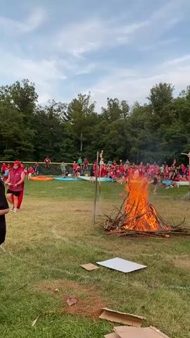 A bonfire burns in a grassy field, surrounded by a large crowd of people. The crowd is mostly comprised of young individuals, many wearing red, blue, or green t-shirts. They are gathered behind a horizontal bar supported by two vertical poles, which is positioned above the bonfire. Two young men stand facing the bonfire, with their backs to the camera, one in a blue tank top and shorts, the other in a blue t-shirt and shorts. Another person is on their hands and knees in front of the bonfire, possibly participating in an activity. In the background, a dense line of trees forms the perimeter of the field, with a hill behind them. The sky is partly cloudy. Scattered around the field are pieces of cardboard and white rectangular objects.