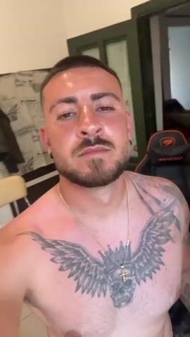 A shirtless man with a short haircut, light beard, and a large tattoo of a winged skull with a crown and cross on his chest is recording a selfie video. He is wearing a gold chain necklace. The camera remains still, focused on his face. Behind him is a partially visible room with a door and a gaming chair.  Electronic music is playing in the background.