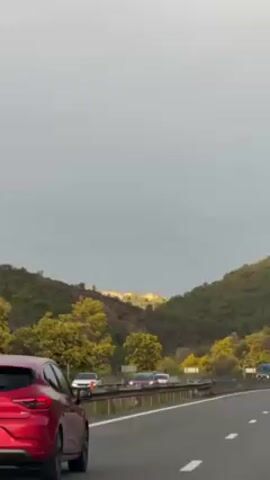 The video shows a red car driving on a highway with hills and trees on the side of the road. The sky is overcast. Other cars are also on the road. The license plate of the red car is visible.