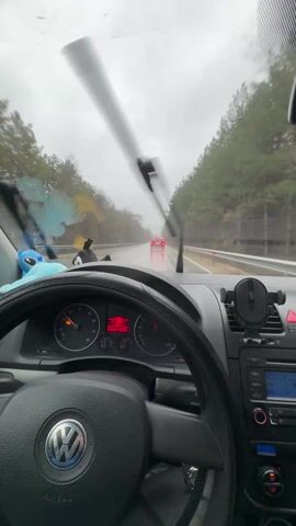 A first-person view from inside a car driving on a wet road. Rain is visible on the windshield, which is being cleared by windshield wipers. The car's dashboard is visible, showing the steering wheel, speedometer, radio, and other controls. There are several small toys hanging from the rearview mirror. A red car is driving ahead. The environment outside the car consists of trees lining the sides of the road. The sky is overcast and grey. The car appears to be moving at a moderate speed.