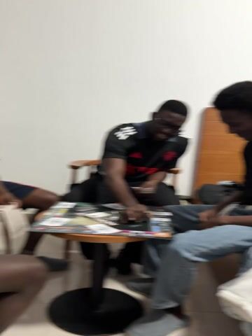 Three individuals are gathered around a small, round table in what appears to be an indoor setting with white walls. One person, wearing a black and red Manchester United jersey, is seated on a wooden chair and appears to be interacting with materials spread out on the table, possibly magazines or books. Another person sits to the right, wearing jeans and a dark top. The third individual is partially visible on the left, showing only legs and part of the seating. The focus is on the person in the jersey and the materials on the table. The scene is somewhat blurry.