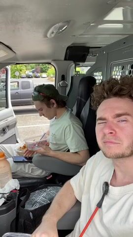 The video shows two young men inside a van. The man in the foreground is in the passenger seat, facing the camera and making a facial expression. He is wearing a light-colored t-shirt. The man in the background is in the driver's seat, facing left and looking down at a clear plastic container of food he is holding. He is wearing a light-colored t-shirt and a green baseball cap. The van's interior is visible, including seats, a door, and a partition behind the driver's seat with circular cutouts. Outside the van, through the open driver's side door, a parking lot with several vehicles and trees can be seen. There are also food items and bags visible in the foreground inside the van.