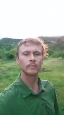 A selfie video shows a fair-skinned man with short blonde hair and a mustache, wearing a green collared shirt. He is standing outside in what appears to be a rural or garden setting. The background features green foliage, including what look like banana trees and some distant hills. The lighting suggests it might be dusk or dawn. The man is looking directly at the camera and the video appears to be recorded with a handheld device as it shows slight movement.