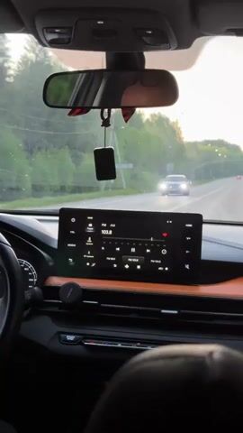 The view from inside a car moving down a road reveals a gray dashboard featuring a large touchscreen displaying a radio interface tuned to 103.8 FM. The interior design includes an orange accent panel. A rearview mirror hangs above with a small black object and a decorative striped piece hanging from it. Through the windshield, a road stretches out with trees lining the sides. The light suggests it is dusk or dawn. Other cars are visible on the road, with one approaching in the opposite lane. The driver's side and steering wheel are partially visible on the left.