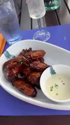 The video shows a table on a patio at what appears to be a resort. On the table are two plates of food, one containing chicken wings with dipping sauce and the other containing fried calamari. There are also a water bottle, a wine glass, an ashtray, and a cell phone on the table. A woman is seated at the table, holding silverware. The patio overlooks a swimming pool and manicured lawn.