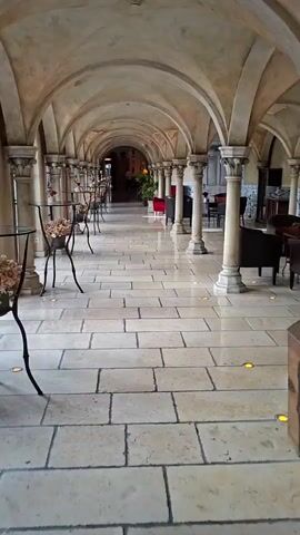 The video shows a long, arched hallway with stone floors and pillars. The hallway is decorated with plants and vases. There are several tables and chairs visible in the background, suggesting this is a public space such as a restaurant or hotel lobby. The lighting is dim, casting long shadows. The camera pans slowly down the hallway, giving a sense of depth and scale. No people are visible in the foreground, but there are figures in the distance at the end of the hallway.
