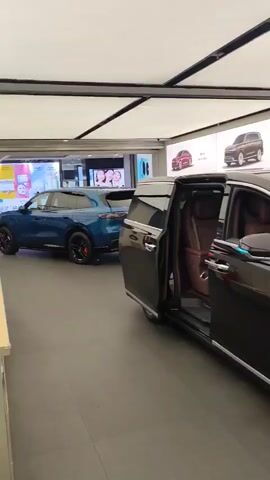 The video pans across a showroom displaying several electric cars. In the foreground, a dark colored luxury sedan with its doors open reveals plush interior seating. To the left, a blue SUV is parked. Further into the showroom, a row of electric cars are displayed, including a red sports car with the designation "智界 Z7" on its license plate, a rose gold sedan, and a maroon sedan. Behind the cars, large digital displays showcase images of various car models. The showroom appears well-lit with a modern, minimalist design, featuring a white panel ceiling.