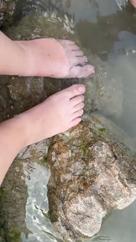 The video opens with a close-up view of two bare feet wading in shallow, clear water. The feet are resting on rocks and sand. The camera then pans out to show a person in the water, further away, with a mountainous, wooded landscape in the background. The water appears to be a lake or a large body of water, with ripples and reflections on the surface. There are some buildings and a red buoy visible in the distance. The lighting suggests it is daytime.