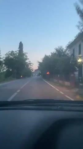 The video shows a first-person view from a car driving on a paved road. The road is bordered by trees on both sides, with some houses visible in the distance. The sky is a clear blue, suggesting it is daytime. There are road signs on the right side of the road, one of which is a yellow triangular warning sign with a red border. The car appears to be moving at a moderate speed. The overall scene is one of a peaceful drive through a rural or suburban area.