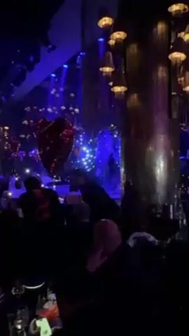 The video shows the interior of a dimly lit venue, possibly a restaurant or nightclub, decorated with various lights and ornaments. Visible are multiple tables occupied by seated individuals. In the foreground, a red heart-shaped balloon floats close to the camera. Further in the background, a stage is illuminated with blue lights, featuring performers in illuminated costumes. A man dressed in black walks casually through the space, smiling and looking towards the performers on the stage. The venue's decor includes large, metallic decorative elements and hanging light fixtures.
