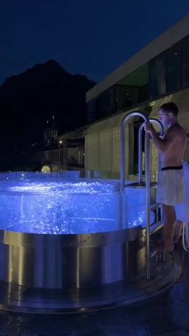 A man in white shorts stands at the edge of a circular, illuminated blue hot tub at night. He is holding onto the silver railing of the steps leading into the tub. Behind him, a building with modern architecture is visible, and in the distance, a dark mountain looms under a night sky. The hot tub is filled with bubbling, illuminated blue water.