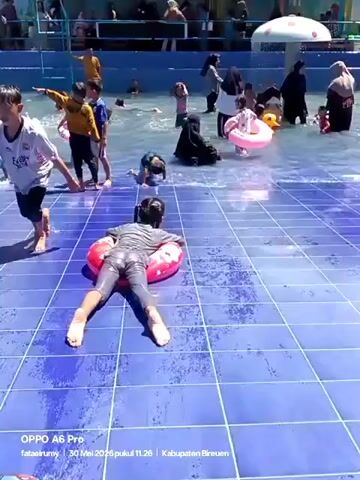 The video shows children and adults enjoying themselves in a water park. The pool has a blue tiled floor and appears to have wave-making capabilities. Many people are in the water, some using inflatable tubes or rings. One child is lying on an inflatable ring on the tiled surface at the edge of the pool. Another child walks across the tiled surface holding an orange inflatable tube. Several other children are standing or walking in the water. In the background, there are structures that appear to be part of the water park facility, including a mushroom-shaped structure. A few adults wearing hijabs are also in the water, supervising the children. The overall atmosphere seems lively and playful.