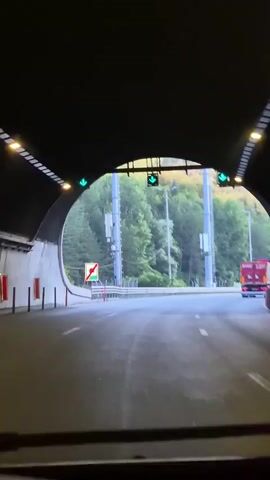 The video captures a view from inside a vehicle emerging from a tunnel onto a highway. The tunnel's entrance is visible in the foreground, with green arrow lights indicating the way forward. Outside the tunnel, the highway curves to the right, bordered by a metal guardrail and a wooded hillside. Several tall, grey structures, likely cell towers, are positioned along the roadside. Further up the hill, three large electrical pylons are visible against a clear sky, suggesting an area with significant power infrastructure. A red truck is driving ahead on the highway. The lighting suggests it is either early morning or late afternoon, with soft sunlight illuminating the scene. The overall setting appears to be a semi-rural or mountainous region. There are no people visible in the frame. A red and white sign with a green border and a red diagonal line across it is visible on the left side of the road just outside the tunnel. The text "Gorno Kamartsi, Bulgaria" indicates the geographical location. The mood is one of transition, moving from an enclosed space to an open one.