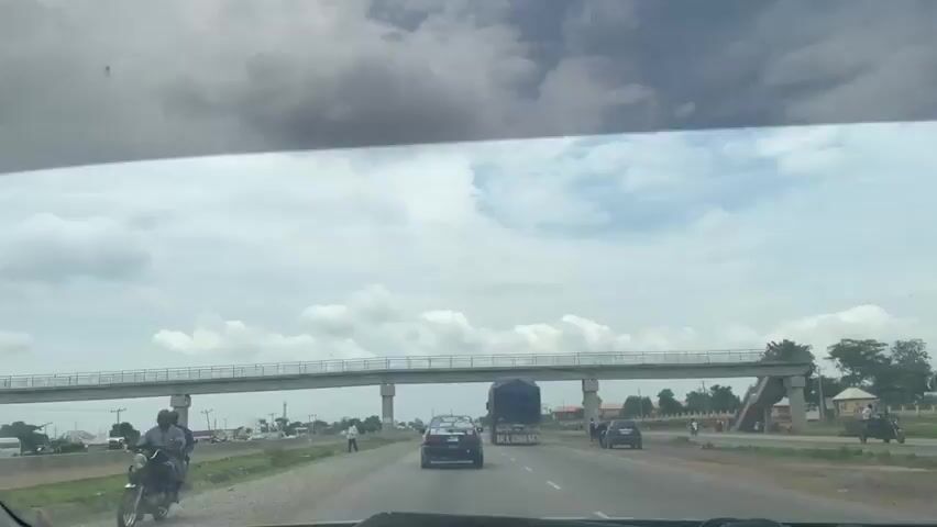 The media file shows a view from inside a vehicle, looking forward through the windshield. The scene is a highway with several cars and a large truck. A pedestrian bridge spans over the highway in the middle distance, supported by concrete pillars. On the right side of the highway, there are some buildings and trees. The sky is cloudy, suggesting it might be overcast or a partially cloudy day. The road appears to be asphalt and there is a shoulder on the left side of the road.

In the foreground, the dashboard of the vehicle is visible. Further ahead, a black sedan is driving in the same lane as the truck. The truck is large, with a dark blue tarp covering its cargo. Some text is visible on the back of the truck, reading "BACK TO JANAH". There are also people visible on the sides of the road, some walking and some standing. The overall impression is a scene of everyday traffic on a highway, possibly in a semi-urban or suburban area, given the presence of both the highway and some structures and trees along the side. The weather appears to be cloudy, and the time of day seems to be daytime. The location context is Gwagwalada, Nigeria.