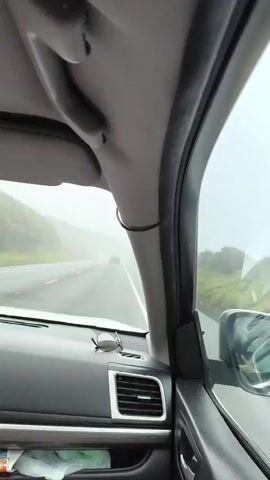 This is a first-person perspective video of someone driving a car on a road that appears to be in a rural or mountainous area. The weather is overcast and foggy, making visibility reduced. The car's interior is visible, including the dashboard, air vent, a pair of sunglasses, and a handle on the ceiling. The road ahead is wet, indicating recent rain or damp conditions. A few other vehicles are visible in the distance on the road. The scenery outside the car includes trees and hills, partially obscured by the fog. The video seems to capture a moment during a road trip or commute in less than ideal weather conditions.