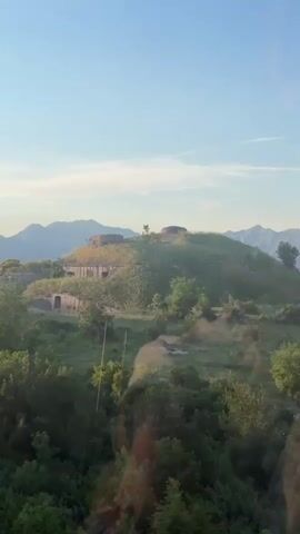The video shows a view from what appears to be a cable car or elevated position, looking out over a green, hilly landscape. In the foreground is dense vegetation with trees and shrubbery. A historic-looking fort or military structure sits atop a hill in the midground. The structure has visible stonework and what appear to be concrete or metal dome-like structures. A range of mountains is visible in the distance, set against a clear, blue sky with a few scattered clouds. The overall light suggests it is either early morning or late afternoon. The camera is relatively still, and there are no visible people or animals.