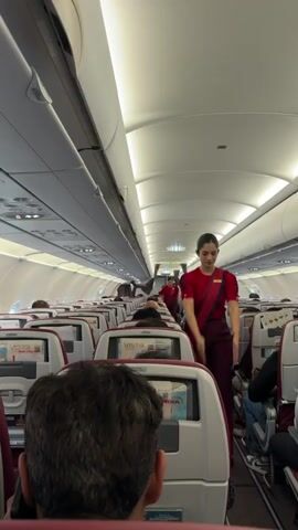 A flight attendant in a red top and maroon pants, with a maroon sash draped over her shoulder, walks down the aisle of a commercial airplane. She is holding a piece of paper and gesturing with her hands. The aisle is lined with rows of seats, many of which are occupied by passengers. The plane's interior is lit by overhead fluorescent lights. In the background, another flight attendant in a similar uniform is visible further down the aisle. The seats have individual entertainment screens. The view is from the perspective of a passenger sitting in one of the seats. The flight attendant appears to be giving safety instructions or information to the passengers.
