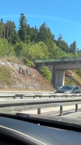 A dashcam view from a car driving on a highway. The camera is positioned low, showing the car's dashboard and windshield. The highway has multiple lanes separated by guardrails and white lines. On the left, a forested hillside with sparse rocks and trees is visible. An overpass crosses the highway. On the right side of the highway, there are other vehicles, including a white bus, a dark car, and a white sedan. The sky is clear and blue, indicating a sunny day. The car is moving at a moderate speed, and the scenery is passing by.