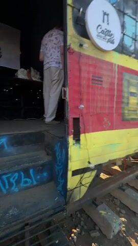 A person wearing a white t-shirt with a pattern and light-colored pants stands facing away from the camera on a wooden platform. To the left, a banner with text and a logo is visible. A table is behind the person, with some items on it. In front of the platform, there are stairs with blue graffiti and an arrow pointing upwards. To the right, a wall is painted with red and yellow stripes, and there is a metal door frame. Further inside, other people are visible, along with green and red lights. One person appears to be holding a microphone. The environment suggests an outdoor or semi-outdoor venue, possibly a stage or performance area.
