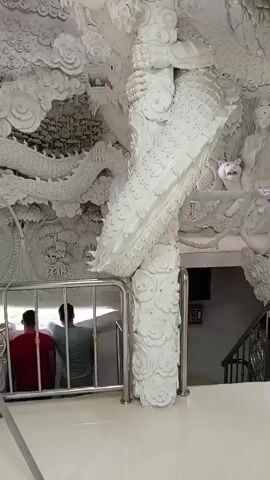 Two individuals, viewed from behind, are standing near a metal railing. The person on the left wears a red shirt, and the person on the right wears a teal shirt. They are facing what appears to be a large, ornate white structure. The structure is heavily decorated with intricate carvings, predominantly featuring dragon motifs and cloud-like patterns. A prominent white dragon sculpture wraps around a pillar. In the background, a staircase with black railings is visible, along with what looks like a framed sign or picture on the wall. The overall environment suggests an indoor space with elaborate, possibly religious or artistic, architectural design.
