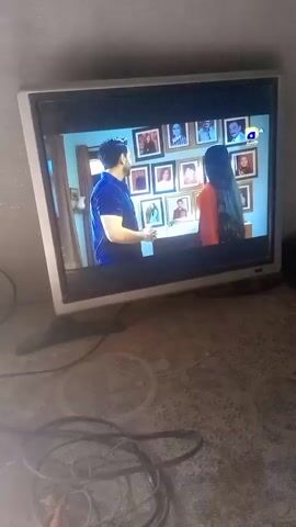 The video shows a silver-framed television displaying a scene from a TV show. Two individuals, a man in a blue shirt and a woman in an orange shirt, are engaged in a conversation. Behind them is a wall adorned with multiple framed photos. The television is placed on a textured surface, with several black cables visible in the foreground.
