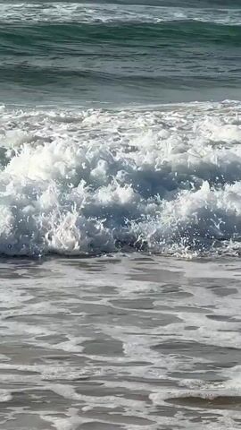 The video shows a close-up of ocean waves crashing on a sandy beach. The water is a deep blue-green, and the waves are white and foamy. The sun is shining, and the water is clear. The scene is peaceful and serene.