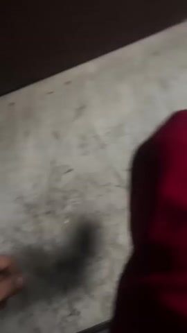 The video captures a close-up view of a person's hand and arm as they reach out towards the ground. The environment appears to be an indoor space with a concrete floor that shows signs of wear and tear, including dirt and scuff marks. A portion of a dark brown surface, possibly furniture or a wall, is visible in the upper right corner of the frame. The lighting is dim, casting shadows and obscuring some details. In the later frames, the camera pans upwards to reveal a light blue wall with what appears to be an electrical outlet and a small, discolored mark. The video ends with a blurry shot of a red object, possibly clothing, in the foreground.