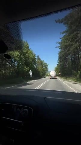 The video shows a car journey with loud music playing. The first shot is from the driver’s perspective, showing a road ahead with a truck. Then, the camera turns towards the passengers. Two people are visible in the front seat, a young woman driving and a young man in the passenger seat. Someone’s hand is visible in the back, holding a glass bottle. The driver covers her eyes momentarily. The camera goes back to the front view, showing the road and truck ahead. The setting appears to be a sunny day, with trees lining the road.