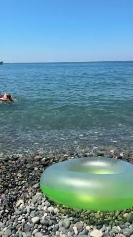 The video shows a bright, sunny day at a pebble beach. In the foreground, a green inflatable ring rests on the stones. The water is blue and clear, with small waves lapping at the shore. Further out in the sea, a group of people are swimming and playing. One person is holding a child while another person is sitting on an inflatable. A ship is visible in the distance on the horizon. The sky is a clear, cloudless blue.