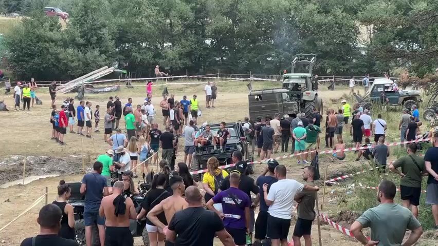 The video shows a large outdoor gathering of people in a rural area. The terrain is uneven and appears to be a dirt field with some muddy patches. There are several vehicles present, including a large green tractor, a military-style truck, and what appears to be an off-road vehicle. A group of people are gathered around the off-road vehicle, and some individuals are sitting in or on it. Other people are standing around in small groups, observing the scene. There are also some elevated structures, possibly ramps or platforms, and some caution tape visible. The background consists of a dense line of trees and a slightly hilly landscape. The overall atmosphere seems to be that of an event or rally, possibly involving vehicles.