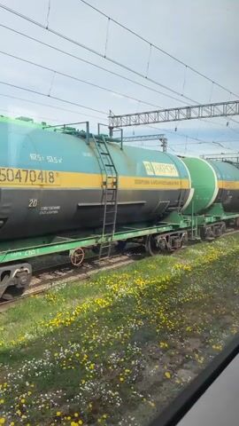 The video shows a train consisting of several tank cars passing by, as viewed from inside another train car. The tank cars are painted in shades of green, blue, and black with yellow horizontal stripes. The logo "АКРОН" is visible on some of the cars along with Russian text. The landscape outside includes green grass and small yellow flowers. Power lines are visible above the trains against a blue sky with light clouds.