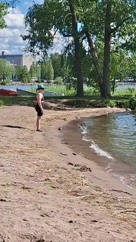 The video shows a sandy beach with shallow, clear water. A person stands on the beach, facing the water, wearing a blue hat and dark shorts. Further out in the water, two people are wading. The scene is lush and green with trees lining the background and a bright blue sky with white clouds above. The sun is shining, indicating a warm, pleasant day. The water is calm with small waves lapping the shore. In the distance, across the water, there are buildings and more trees, suggesting a populated area or park. The overall mood is relaxed and peaceful, typical of a summer day at the beach. The time of day appears to be daytime, likely mid-morning or afternoon, given the bright sunlight. There is no visible text in the video.