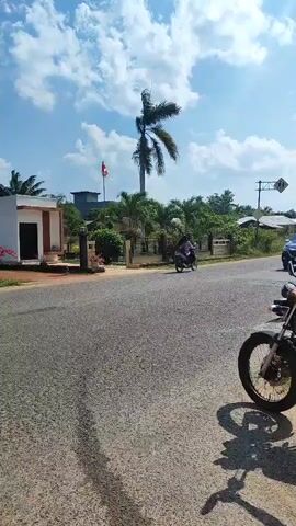 The video shows an outdoor scene with a road and a sidewalk. On the sidewalk, there are two motorcycles. One is parked in the foreground, with its front wheel visible. The other motorcycle is ridden by a person wearing a dark t-shirt and blue shorts, who is facing away from the camera. Further down the road, a dark-colored SUV is driving away from the camera, and another motorcycle with a rider is visible behind it. In the background, there is a building with an Indonesian flag flying on a flagpole, a tall palm tree, and other vegetation. The sky is blue with white clouds. The overall lighting suggests it is daytime.
