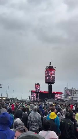 A wide shot shows a very large crowd of people gathered outdoors at what appears to be a concert or festival. Many individuals in the densely packed crowd are wearing rain gear such as ponchos and hooded jackets. They are all facing towards multiple large video screens in the distance that display images of performers on a stage. Tall structures support these screens and other equipment. A large building with some visible text, possibly "PINK," is in the background behind the stage area. The sky is heavily overcast with gray clouds.
