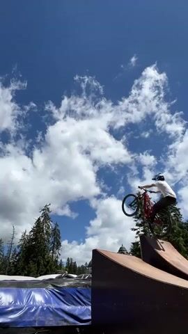 A mountain biker performs a trick in mid-air against a blue sky with scattered clouds. The biker is wearing a white t-shirt, black shorts, and a helmet. The biker's red bicycle is also in the air. Below the biker are two ramps, one made of wood and the other covered in blue tarp. Tall evergreen trees are visible in the background. The scene appears to be outdoors on a sunny day.