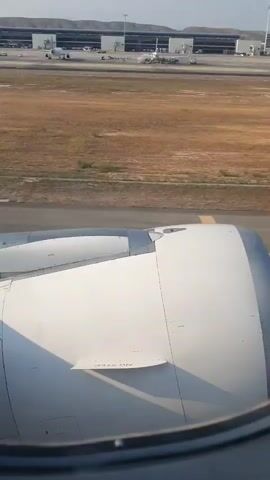 This is a first-person view from an airplane window during takeoff. The camera is looking out the window at the airplane's engine and wing. In the distance, an airport terminal building and runways are visible under a clear sky. The airplane is on the runway, and the engine is clearly visible. The scene appears to be set during the day, with bright sunlight casting shadows. The video is short, lasting only a few seconds, and depicts the initial moments of takeoff.